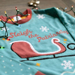 Load image into Gallery viewer, A flat lay of the Sleigh The Patriarchy Holiday Sweater. There are colorful holiday lights, bows, and ornaments around them.