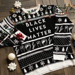 Load image into Gallery viewer, A flat lay of the Black Lives Matter Holiday Sweater. There are colorful holiday lights, ornaments, and bows around it.