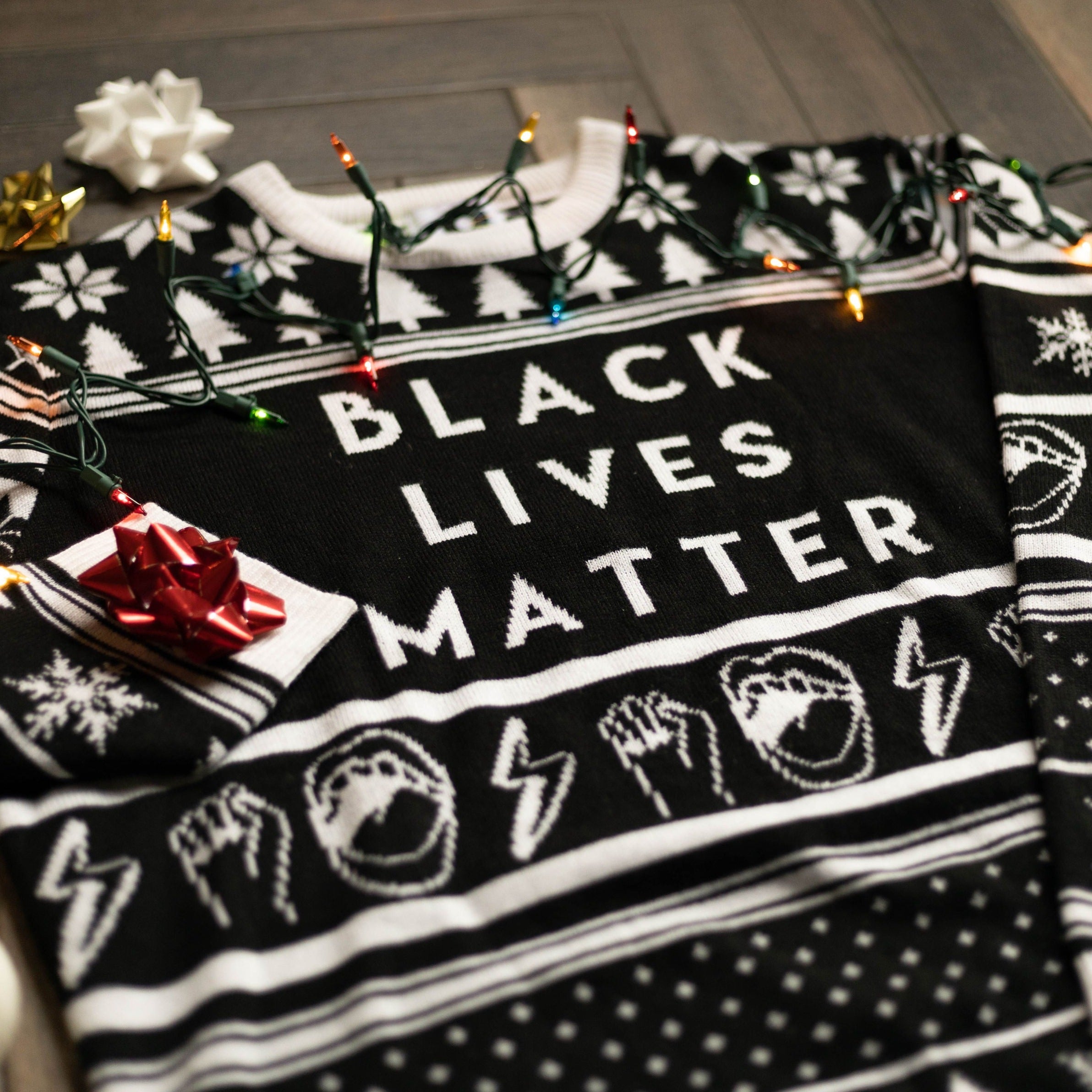 A flat lay of the Black Lives Matter Holiday Sweater. There are colorful holiday lights, ornaments, and bows around it.