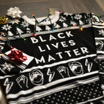 Load image into Gallery viewer, A flat lay of the Black Lives Matter Holiday Sweater. There are colorful holiday lights, ornaments, and bows around it.