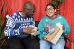 Load image into Gallery viewer, A person wearing the Get In Good Trouble Holiday Sweater holding a gift. They are sitting next to someone wearing the Sleigh The Patriarchy Holiday Sweater who is also holing a gift. There are plants with ornaments on them and a red background.