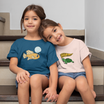 Load image into Gallery viewer, girls sit on the stairs holding hands, one in a teal shirt with "climate justice is fantastic" design and one in a peach "feminism is turtley awesome" design