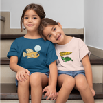 Load image into Gallery viewer, girls sit on the stairs holding hands, one in a teal shirt with "climate justice is fantastic" design and one in a peach "feminism is turtley awesome" design
