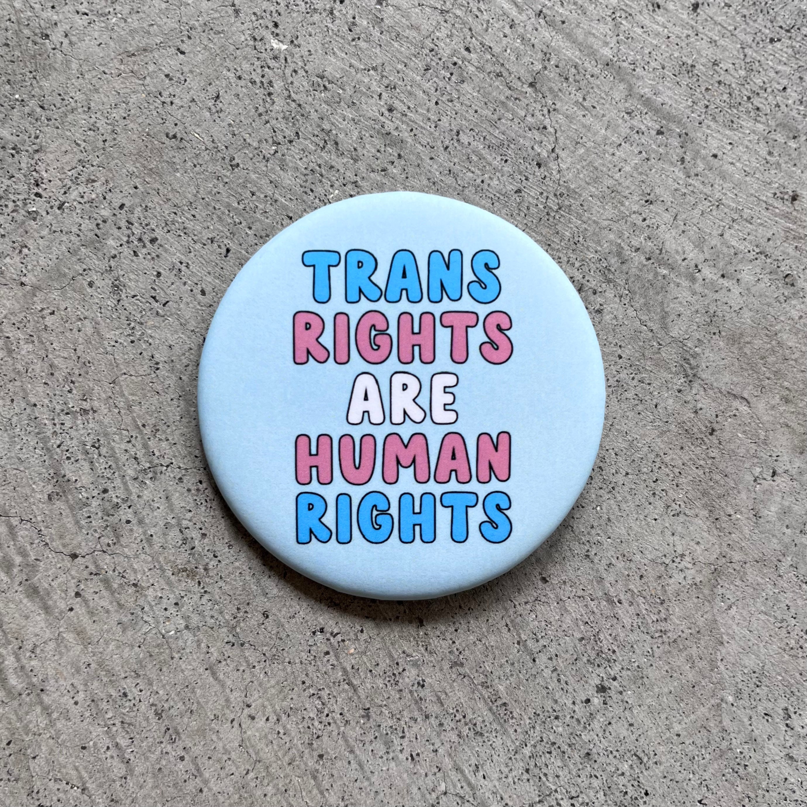Round, Light Blue button with "Trans Rights Are Human Rights" in the colors of the Trans flag