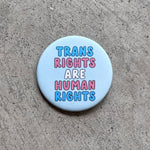 Load image into Gallery viewer, Round, Light Blue button with &quot;Trans Rights Are Human Rights&quot; in the colors of the Trans flag
