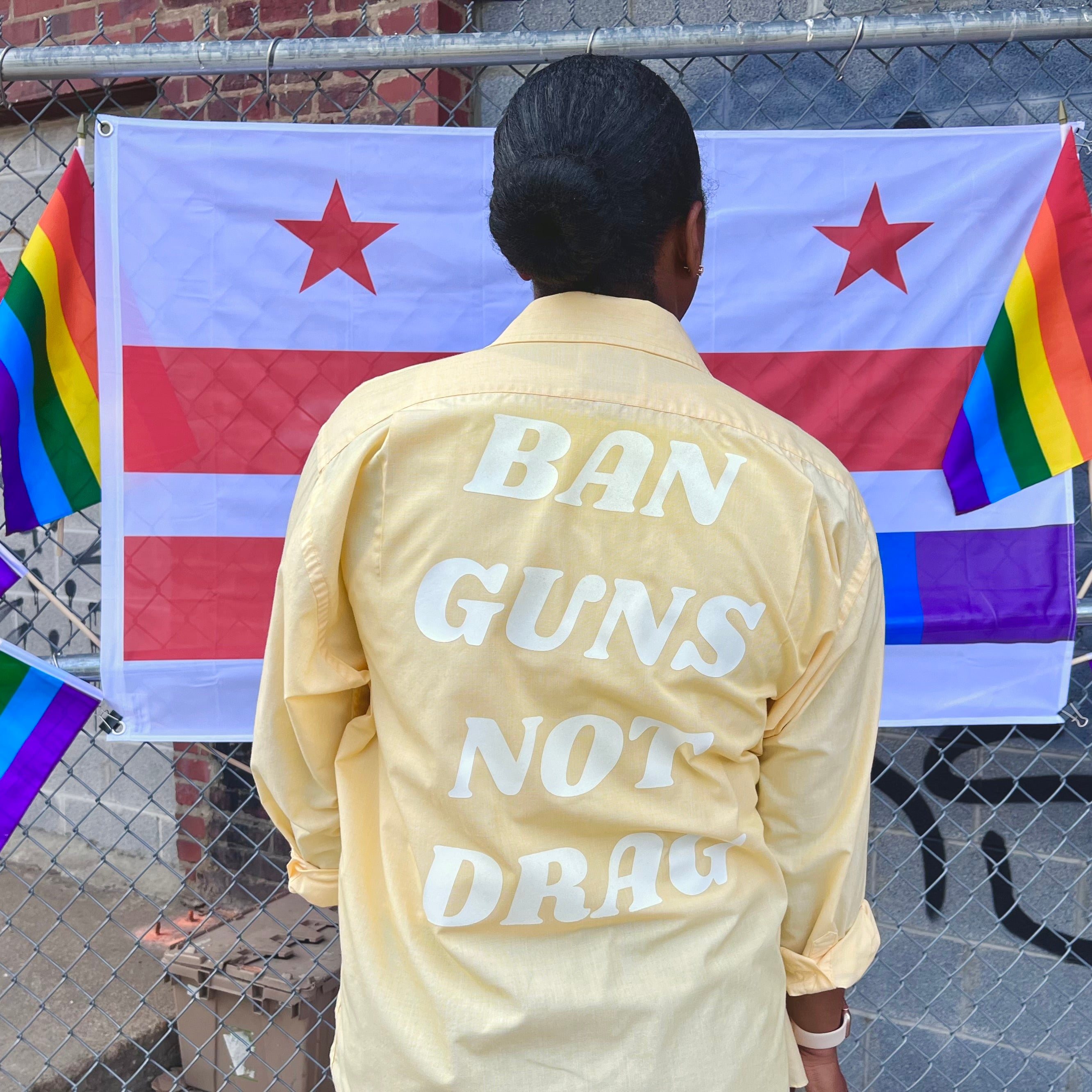 Woman in front of rainbow flags wears a yellow upcycled button down with "Ban Guns Not Drag" printed across the back in a 70s font.