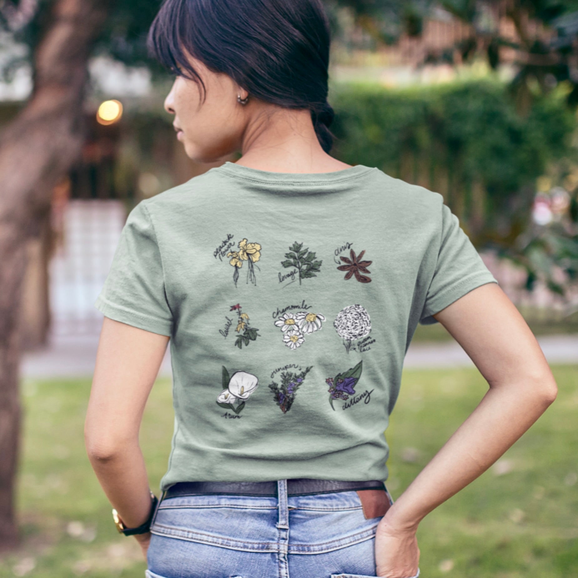 Person wearing a slightly weathered sage green unisex tee facing away from us. The back of the tee features a design of herbs and plants that can be used to self manage an abortion