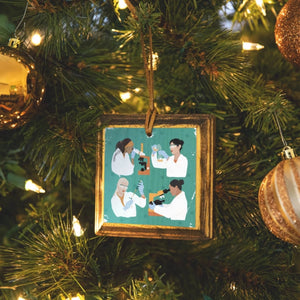 Square wooden ornament with four diverse women in lab coats with scientific equipment on a teal background