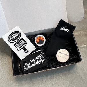 Gift box including: black socks with "Resist" design, Policy and change button, Our Home is on Fire button, coin purse with "Keep the Coins, I want Change!" design, and We all quit Capitalism sticker