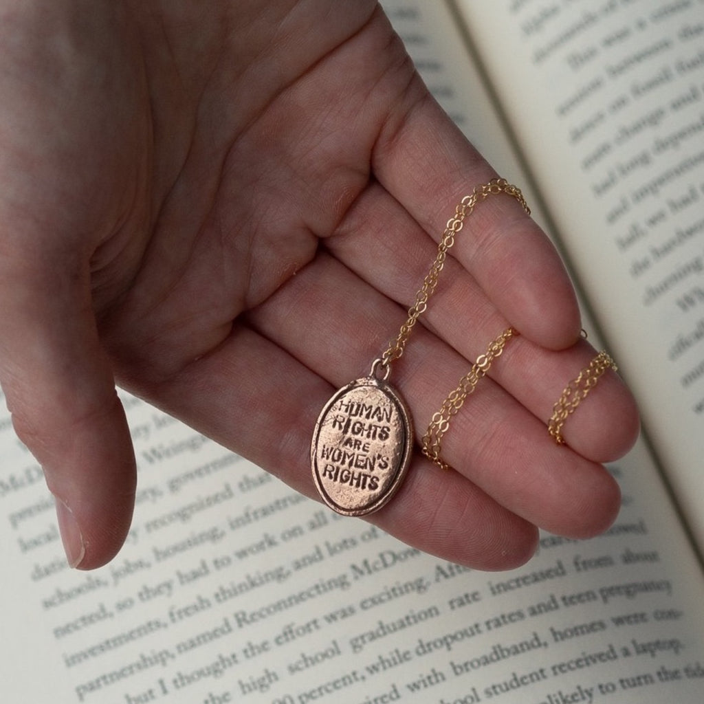 "Human Rights are Women's rights" HRC quote bronze medallion necklace sitting in a hand above a book 