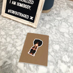 Load image into Gallery viewer, Photo of the sisterhood sticker on a plain notebook next to a sign.
