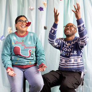A person wearing the Sleigh The Patriarchy Holiday Sweater. Next to them is a person wearing the Get In Good Trouble Holiday Sweater, they are throwing bows in the air that are falling around them.