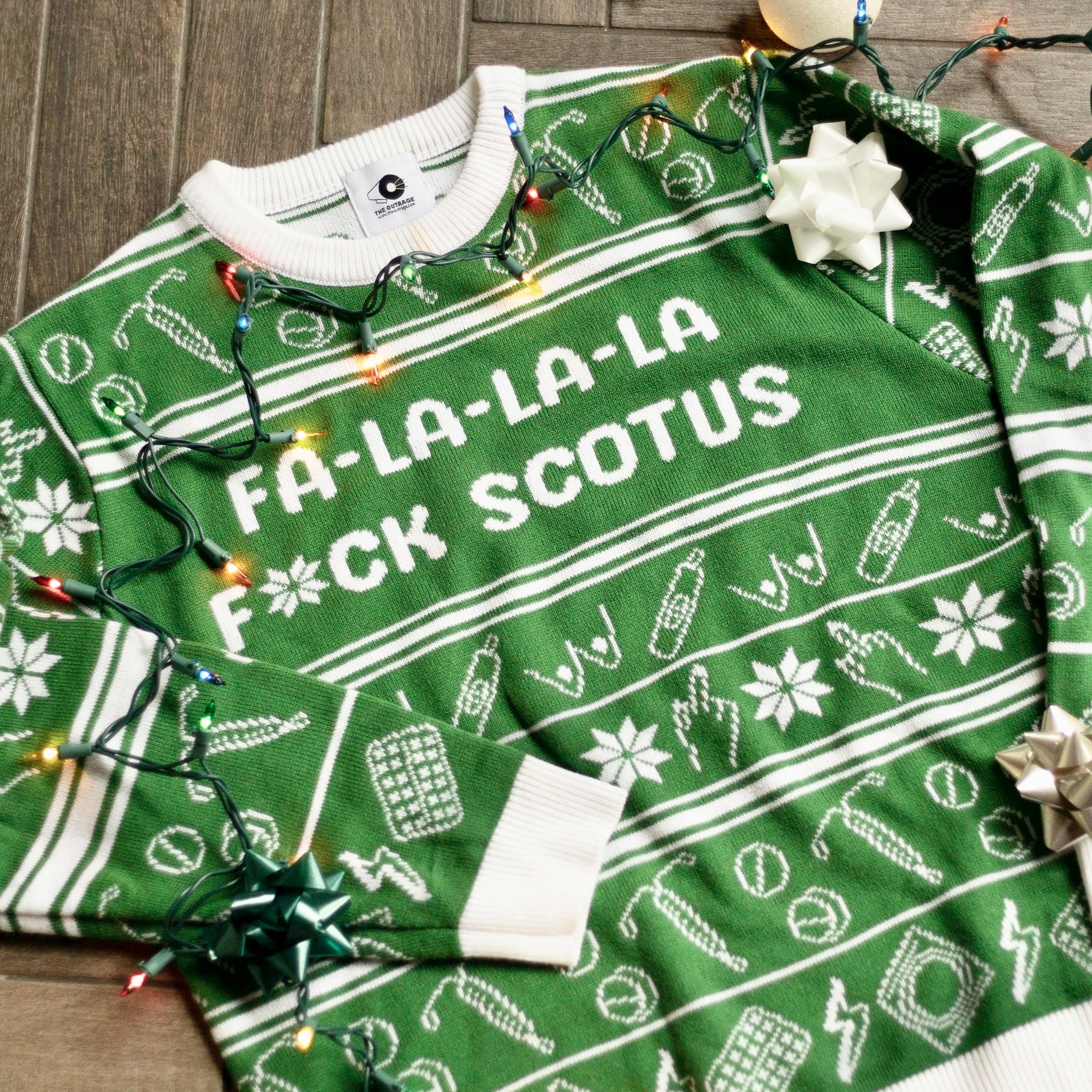 Flat lay of the Fa-La-La-La F*ck SCOTUS Holiday Sweater. There are colorful holiday lights on it and bows and ornaments around it.