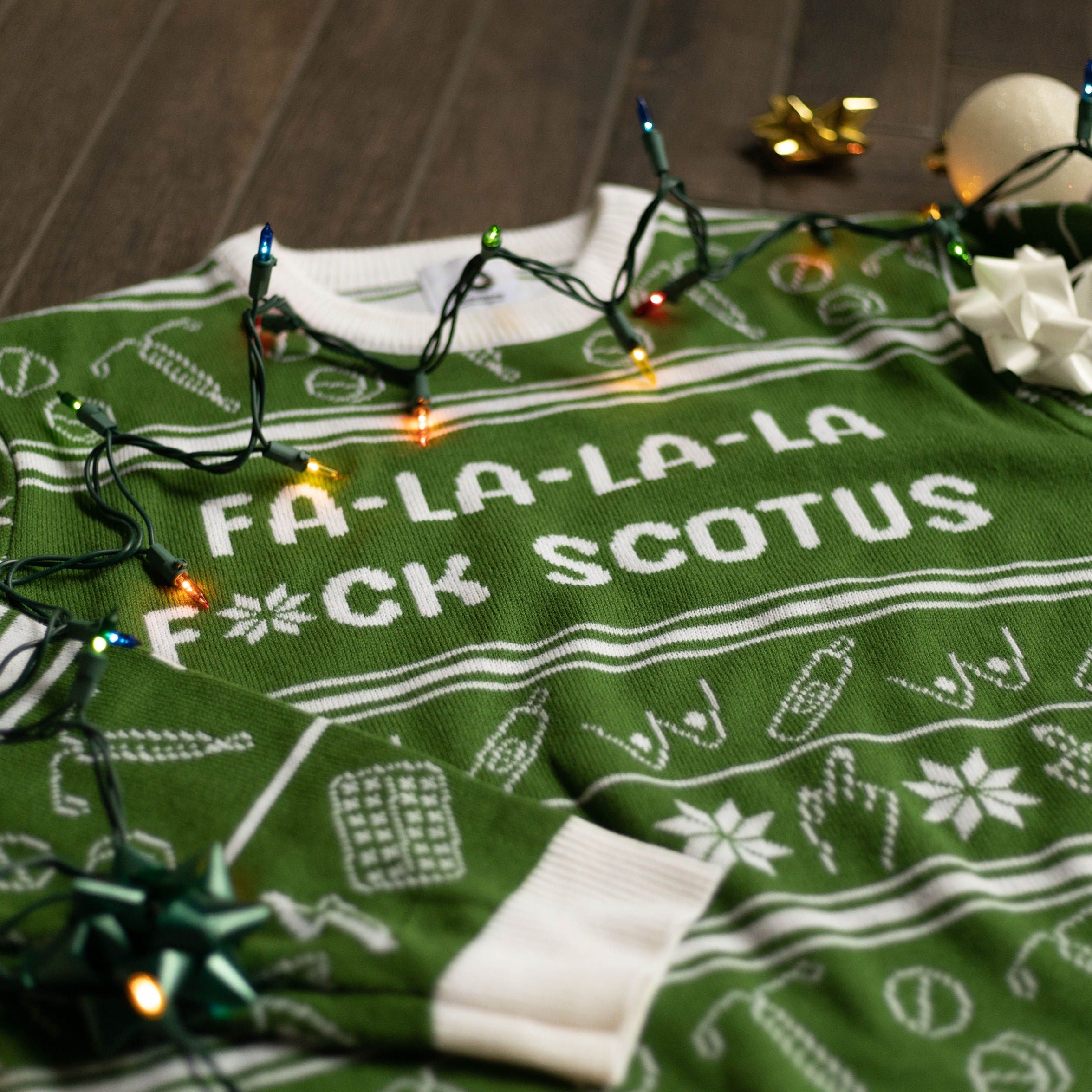 Flat lay of the Fa-La-La-La F*ck SCOTUS Holiday Sweater. There are colorful holiday lights on it and bows and ornaments around it.