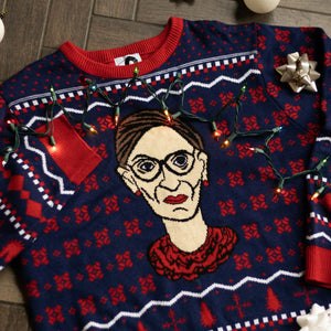 A flat lay of the RBG Holiday Sweater. There are colorful holiday lights, bows, and ornaments around it.