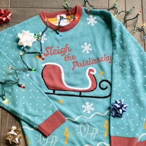 A flat lay of the Sleigh The Patriarchy Holiday Sweater. There are colorful holiday lights, bows, and ornaments around them.