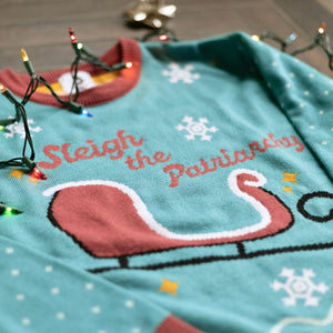 A flat lay of the Sleigh The Patriarchy Holiday Sweater. There are colorful holiday lights, bows, and ornaments around them.