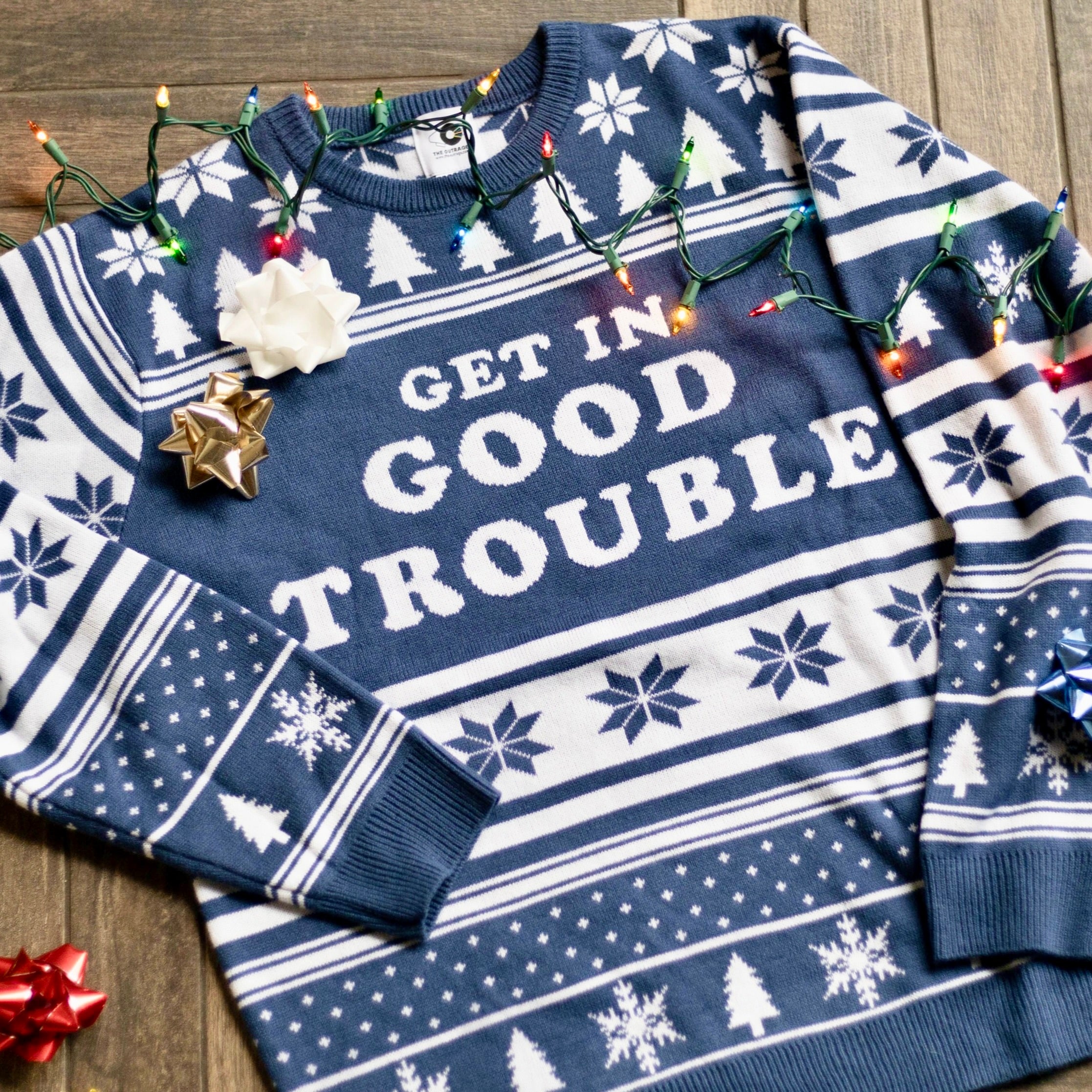 A flat lay of the Get In Good Trouble Holiday Sweater. There are colorful holiday lights, ornaments, and bows around it.