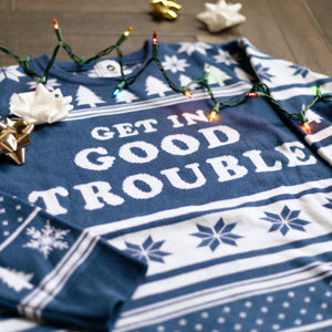A flat lay of the Get In Good Trouble Holiday Sweater. There are colorful holiday lights, ornaments, and bows around it.