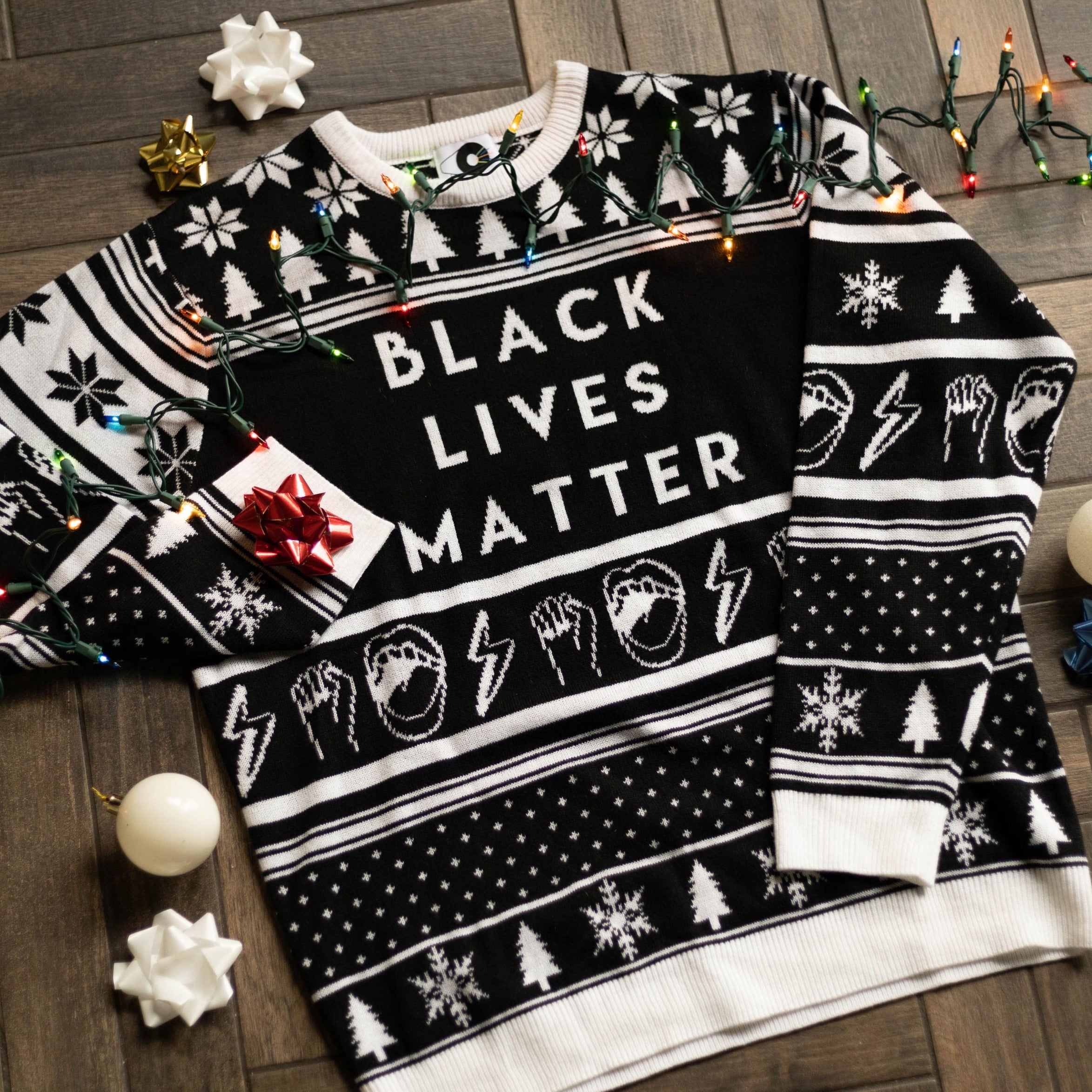 A flat lay of the Black Lives Matter Holiday Sweater. There are colorful holiday lights, ornaments, and bows around it.
