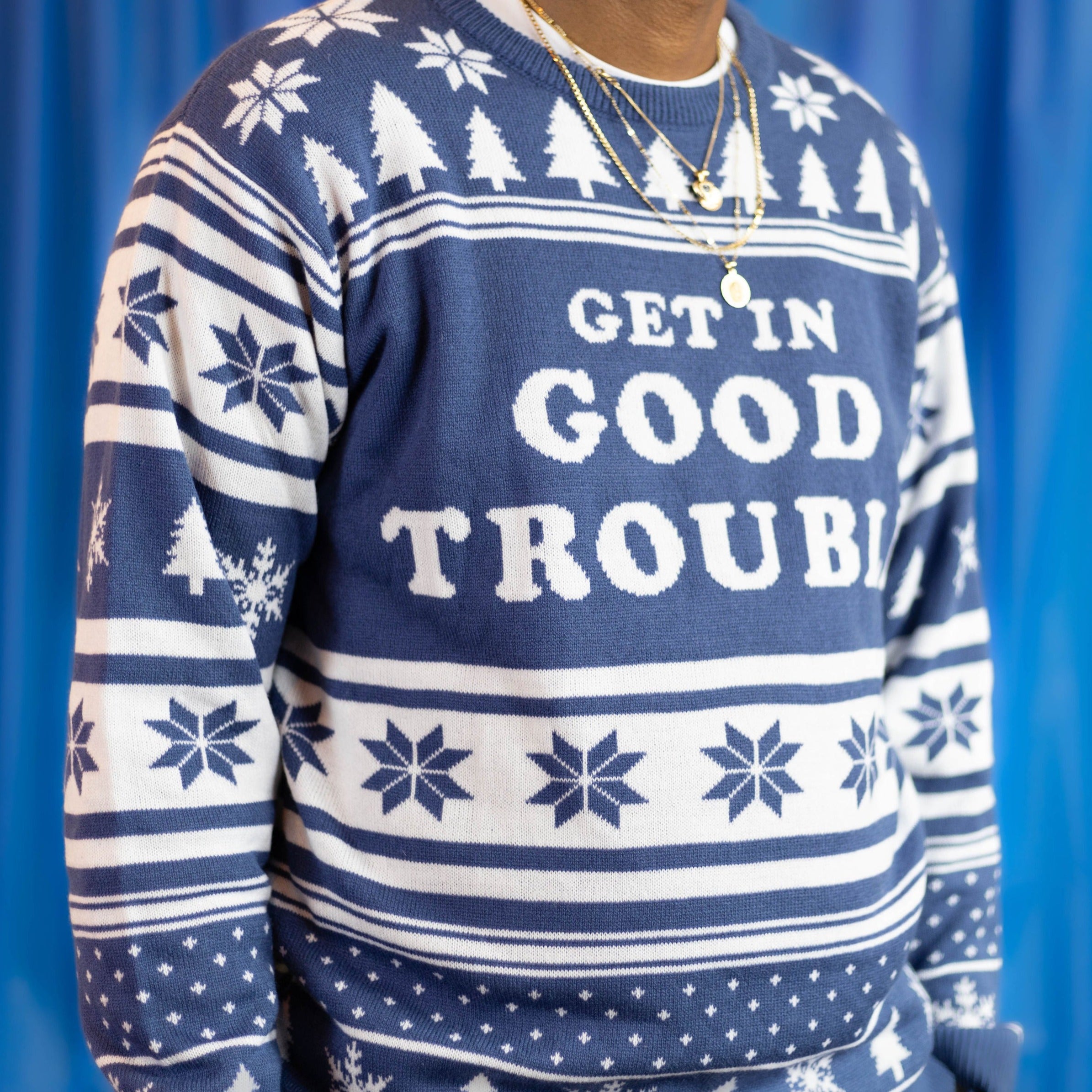 A person wearing the Get In Good Trouble Holiday Sweater. There is a blue background.