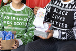 Load image into Gallery viewer, A person wearing the Fa-La-La-La F*ck SCOTUS Holiday Sweater holding a gift. Sitting next to them is a person wearing the Black Lives Matter Holiday Sweater holding a gift. There is a red background.
