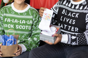 A person wearing the Fa-La-La-La F*ck SCOTUS Holiday Sweater holding a gift. Sitting next to them is a person wearing the Black Lives Matter Holiday Sweater holding a gift. There is a red background.
