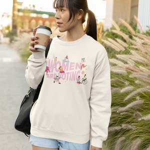 Young woman with dark hair, bangs, and coffee cup wears a cream crewneck sweatshirt with "Women Are Voting" design in pink surrounded by inclusive cartoon femme figures.