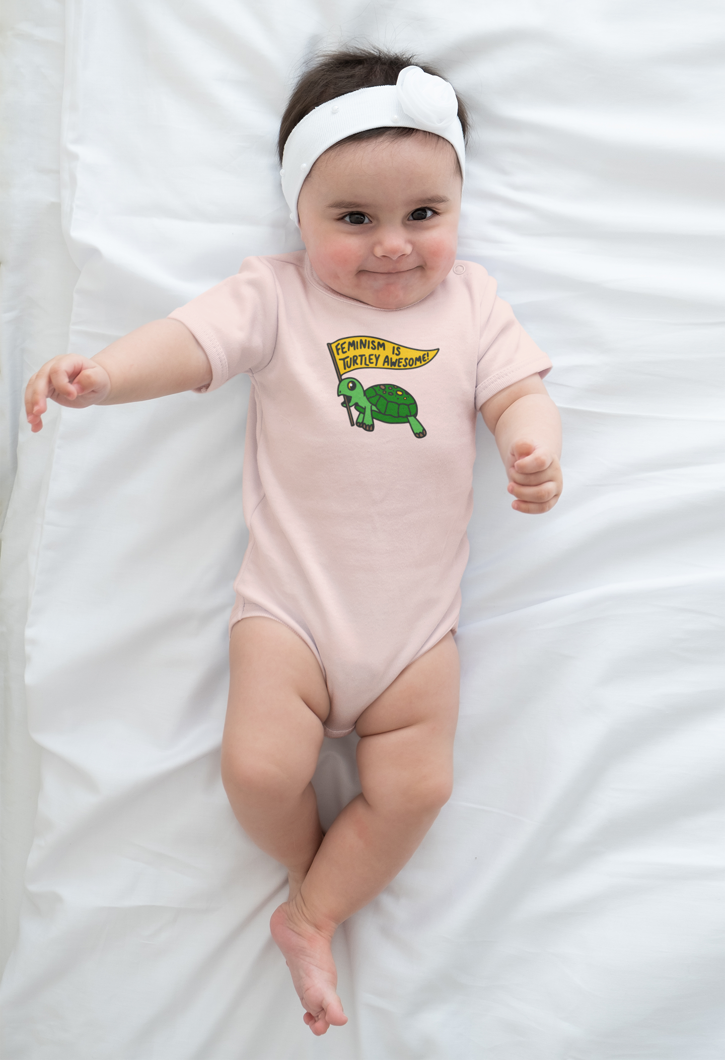 baby with headband wearing peach shirt with "Feminism is Turtley Awesome" onesie