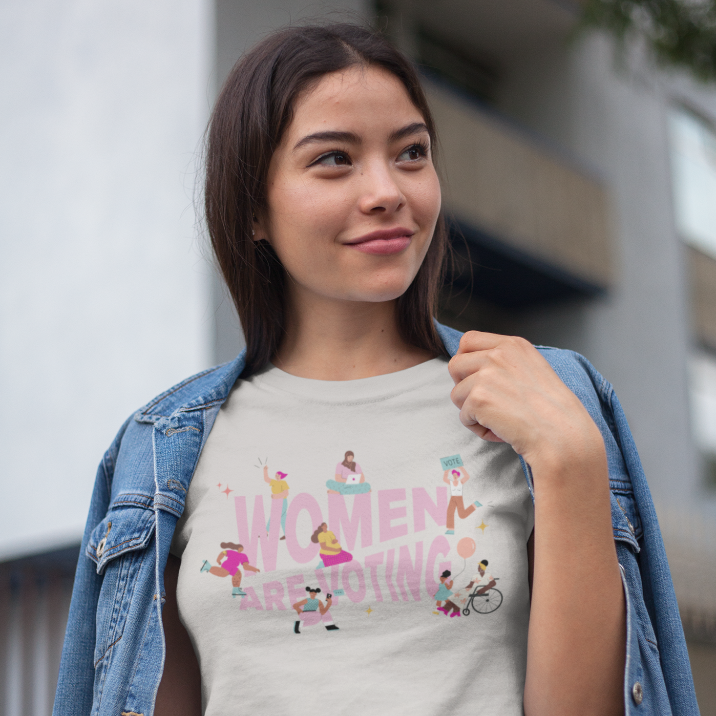 A young woman with dark hair and a jean jacket over her shoulders wears a cream t-shirt with the phrase "Women Are Voting" in pink surrounded by inclusive femme cartoon figures
