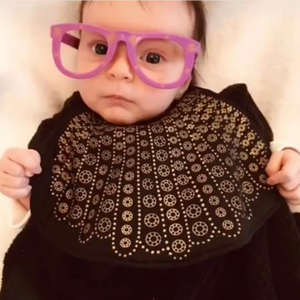 Baby wearing purple glasses and a bib with RBG dissent collar pattern